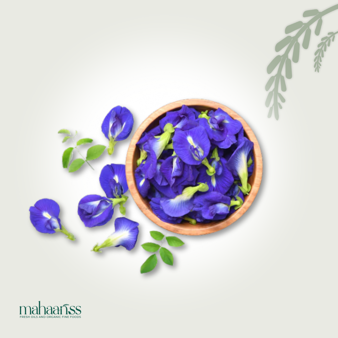 Bowl of blue butterfly pea flowers with green leaves on a light gray background, featuring the brand 'maharishi'.